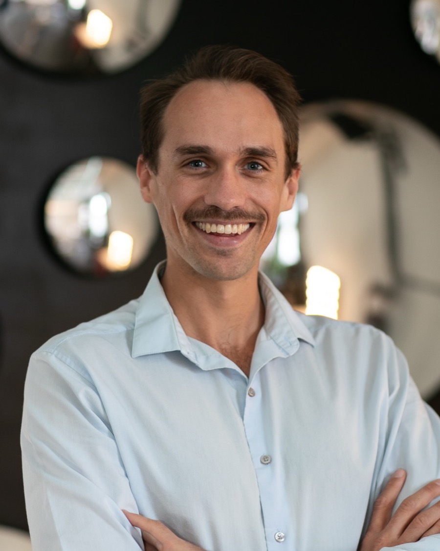 Benza Maman smiling in a light blue shirt, arms crossed, in front of round mirrors